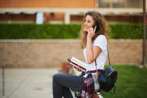 Female srudent talking on the smart phone and holding books while sitting on the bench. photo