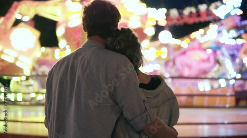 Wallpaper Mural Young couple visiting an amusement park arcade together and hugging while standing next to a carousel ride with lights at night. Standing from the back Torontodigital.ca