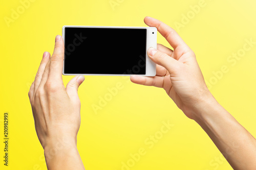 Mockup of female hands holding white frameless cellphone with black screen and making selfie at isolated yellow background.