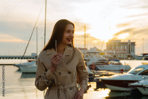 Foto beautiful long-haired smiling girl in trendy beige coat posing in seaport at sun
