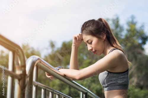 Young woman relaxing after jogging exercise on fence at park to freshen her body and enjoy warm light in morning. Young asian woman take a break from running execise. Outdoor exercise activity. © tirachard