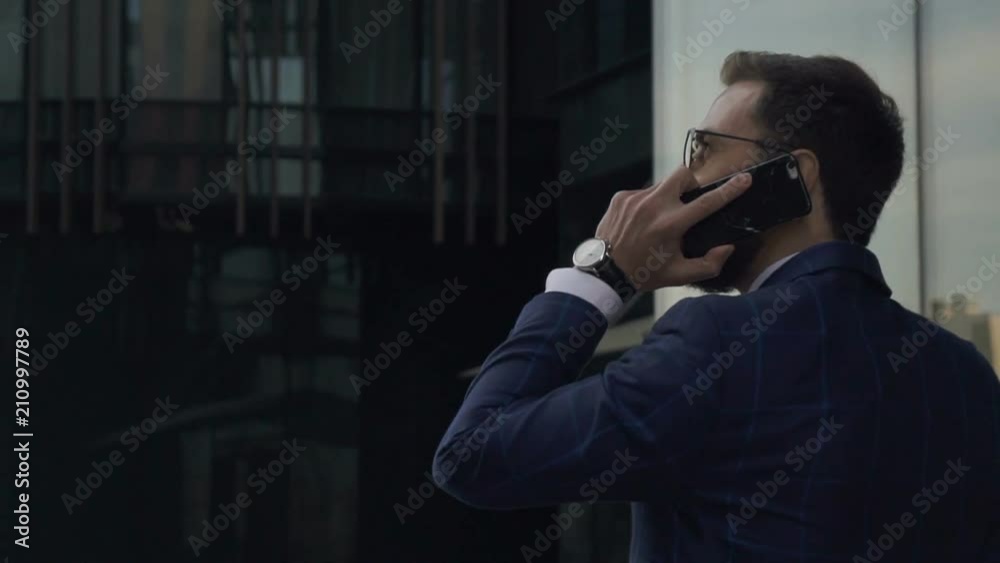 Young handsome businessman is talking on phone walking along New York street, back view of elegant man in suit is in business conversation, going on background of cityscape. Concept: mobile calling