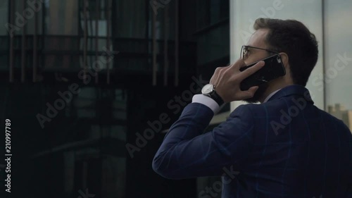 Young handsome businessman is talking on phone walking along New York street, back view of elegant man in suit is in business conversation, going on background of cityscape. Concept: mobile calling