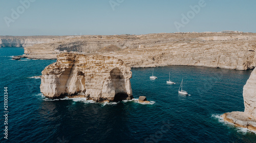 Aerial view of three white yacht sailing boats in the bay of deep blue mediterranean sea with rocky cliffs and island in Gozo, Malta. Shot from above. Travel concept photo