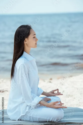 Obraz side view of young asian woman in ardha padmasana (half lotus pose) on yoga mat by sea