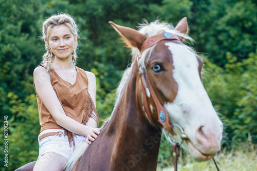 People and animals friendship, hippotherapy concept. Portrait of happy pensive woman cowgirl, riding a brown horse. Clothed white jeans shorts, brown leather vest. Has slim sport body.