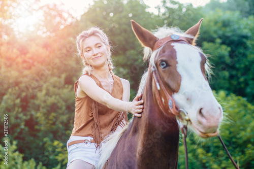 People and animals friendship, hippotherapy concept. Portrait of happy pensive woman cowgirl, caress a brown horse while riding. Clothed white jeans shorts, brown leather vest. Has slim sport body.
