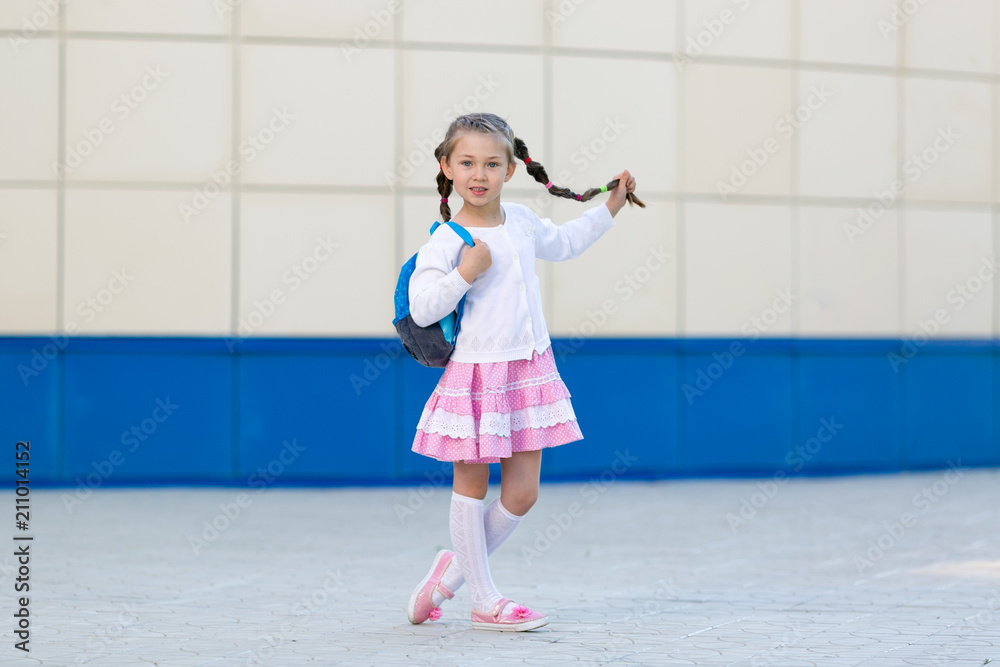 The girl is a young fashionista posing. A little beautiful girl in a summer dress with a backpack is standing by the wall in the city. The girl is a young fashionista posing. A little beautiful girl in a summer dress with a backpack is standing by the wall in the city.