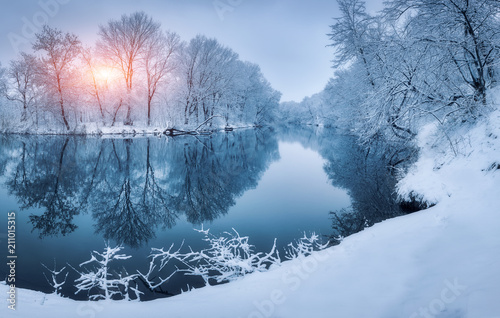 Winter forest on the river at sunset. Colorful landscape with snowy trees, frozen river with reflection in water. Seasonal. Snow covered trees, lake, sun and blue sky. Beautiful forest in snowy winter