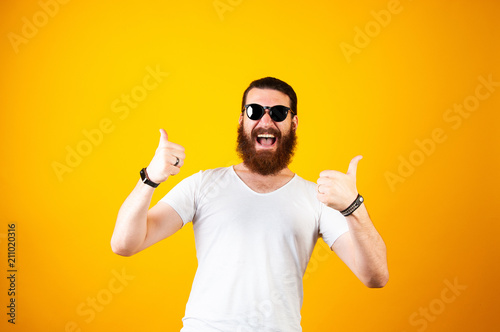 Фотография Excited young hipster bearded man wearing glasses and a white T-shirt laughing and smiling at the camera isolated on a yellow background