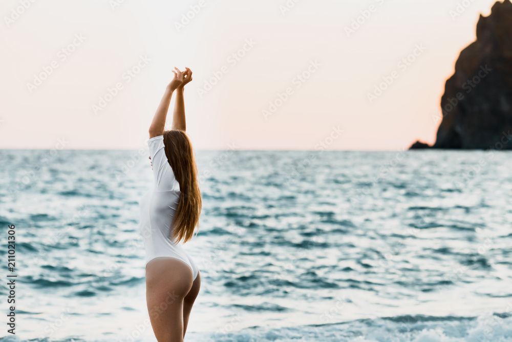 Beautiful cute young girl with long brown hair and white swimsuit is standing a half-face on the beach. Blurred blue water of the sea at summer sunny day. Copy space