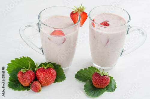 Two transparent glasses with milk strawberry cocktail on a white wooden table. Next to strawberry berries with leaves.