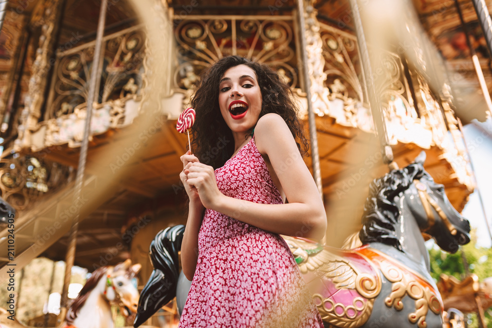 Young joyful lady with dark curly hair in dress standing with lolly pop candy in hands and happily looking in camera with beautiful carousel on background