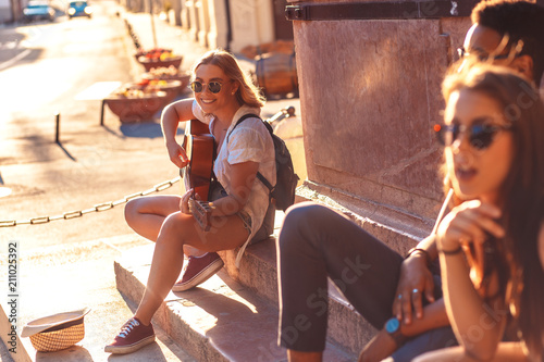 Group of friends having good time at the city street. Girl playing guitar.