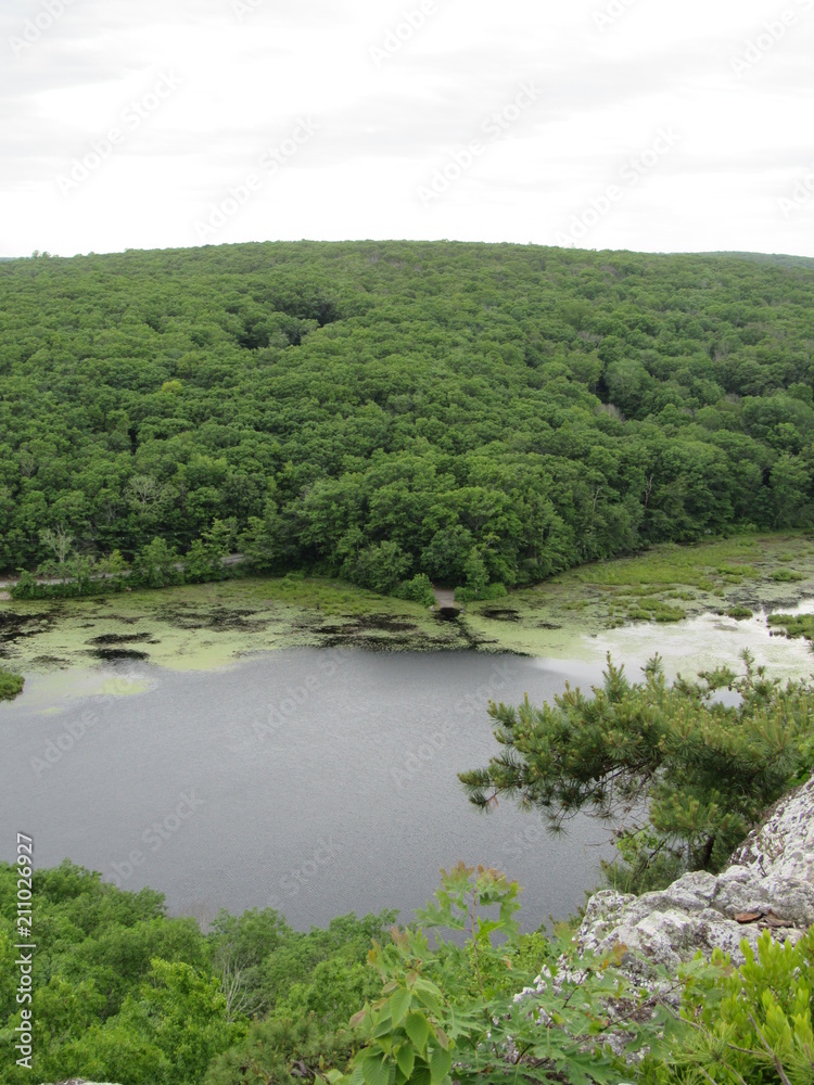 View over the edge of a cliff on a mountain on Lantern Hill trail in Connecticut, with a pond below in the distance View over the edge of a cliff on a mountain on Lantern Hill trail in Connecticut, with a pond below in the distance