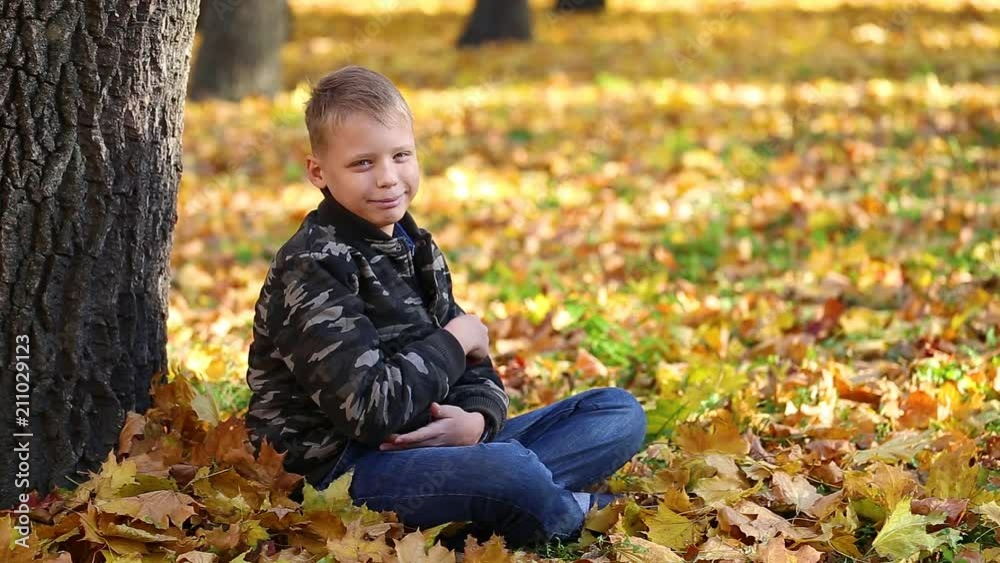 Portrait of cute funny white boy looking at camera smiling while sitting alone on ground in park covered with bright orange maple leaves. Kid shows thumb up as sign of success or likeness. Portrait of cute funny white boy looking at camera smiling while sitting alone on ground in park covered with bright orange maple leaves. Kid shows thumb up as sign of success or likeness.