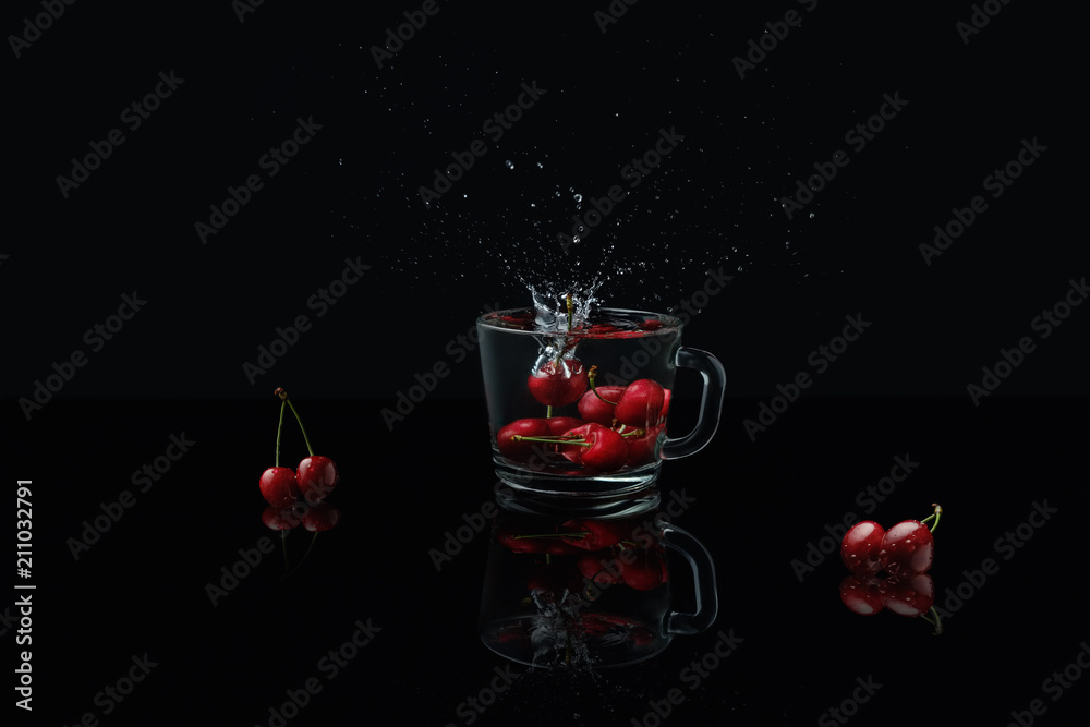 Macro photo of a clear glass mug with cherries in front of a black background on a black table made of glass. Reflection of a glass mug with cherries. Falling berries create splashes of water.