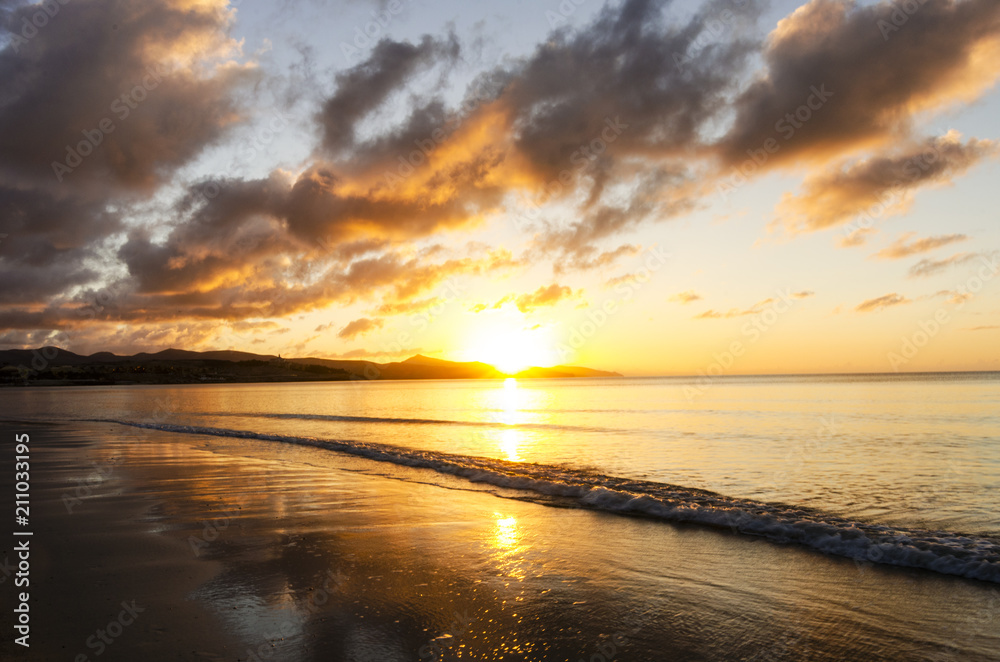 Yellow and orange sunrise on the beach with sun reflection on the wet sand and small wave on the calm ocean surface