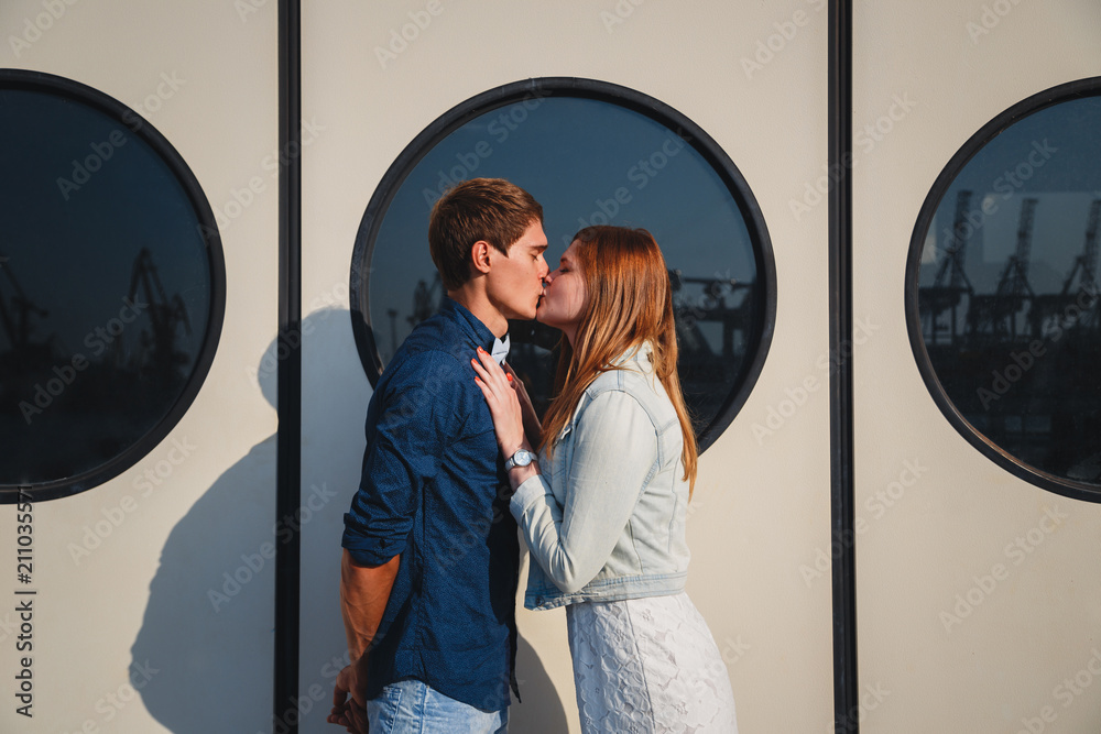 Cute young beautiful hipster couple kissing at port, on the white wall