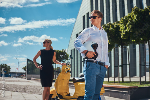 Attractive couple - handsome stylish guy dressed in a white shirt and jeans and charming blonde woman wearing black dress standing near yellow classic Italian scooter against a skyscraper. Vacation © Fxquadro