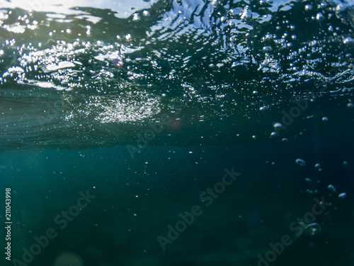 Sea water wave with bubbles closeup underwater photo. Blue sea surface from below. Ripples on sea surface.