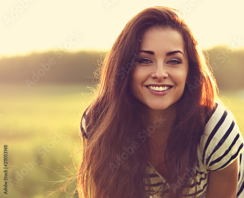 Beautiful toothy smiling young woman looking happy with long amazing bright hair on nature bright sunset summer background. Closeup toned color portrait