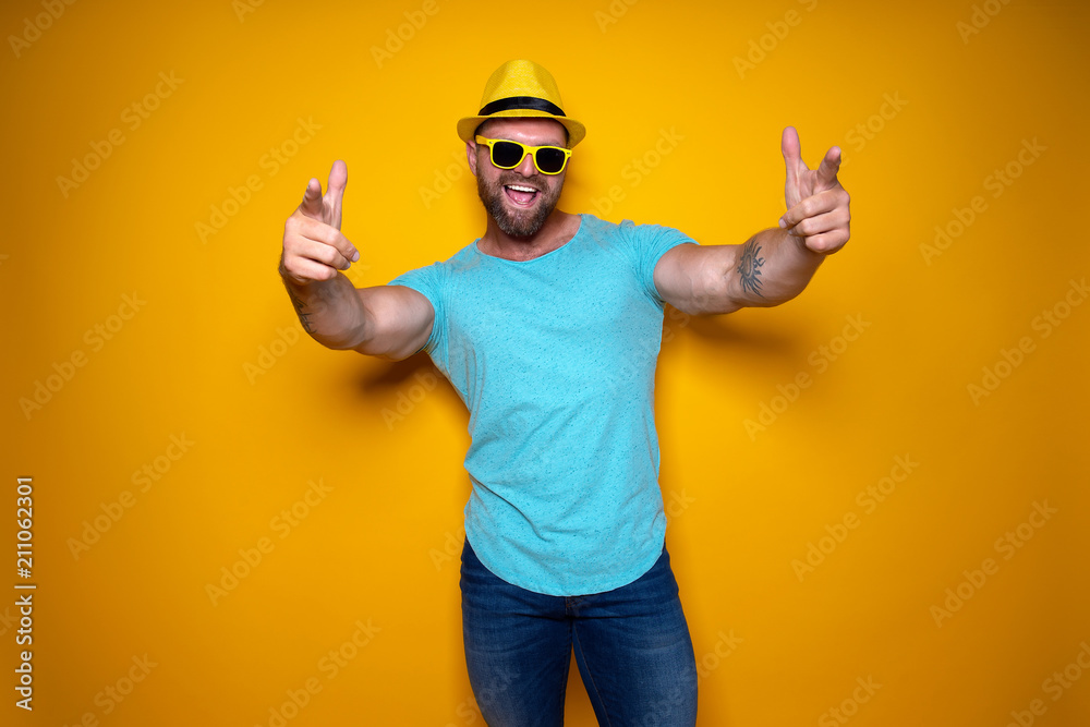 Happy male in blue shirt, sunglasses and yellow hat celebrating success standing with open mouth smiling and pointing fingers of stretched out arms forward on yellow background