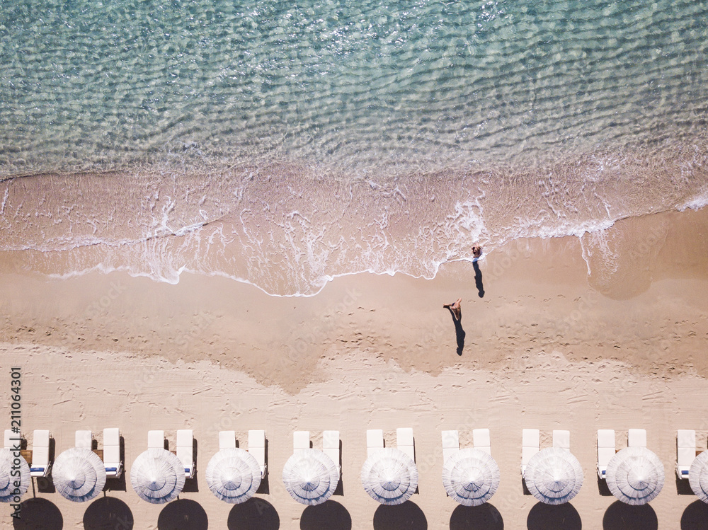 Aerial view of amazing turquoise sea with white umbrellas and sun loungers. Two people are walking on the shore. Beautiful sunny summer day in Sardinia, Mediterranean sea, Italy..