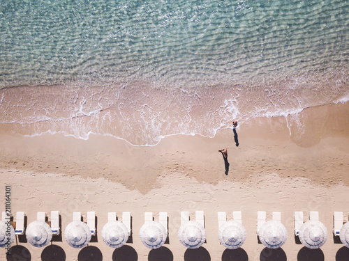 Obraz na płótnie Aerial view of amazing turquoise sea with white umbrellas and sun loungers. Two people are walking on the shore. Beautiful sunny summer day in Sardinia, Mediterranean sea, Italy.. w salonie