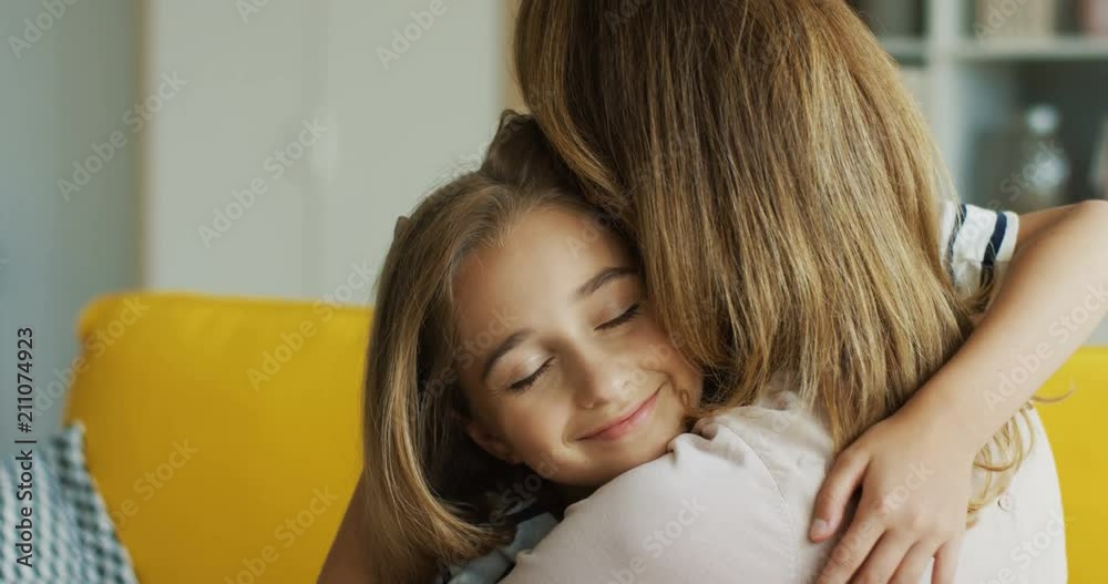 Portrait of the cute blonde teenage girl embracing her mother and smiling nicely in the living room. Close up. Inside. Portrait of the cute blonde teenage girl embracing her mother and smiling nicely in the living room. Close up. Inside.