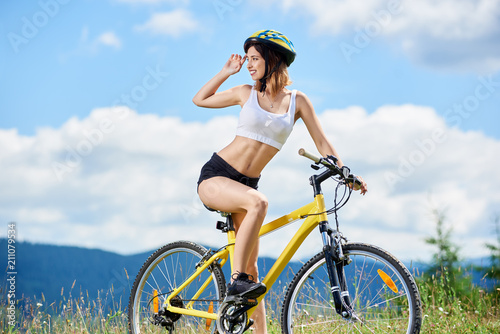 Beautiful sporty woman cyclist posing on yellow bicycle, wearing helmet, enjoying sunny day in the mountains against blue sky with clouds. Outdoor sport activity, lifestyle concept