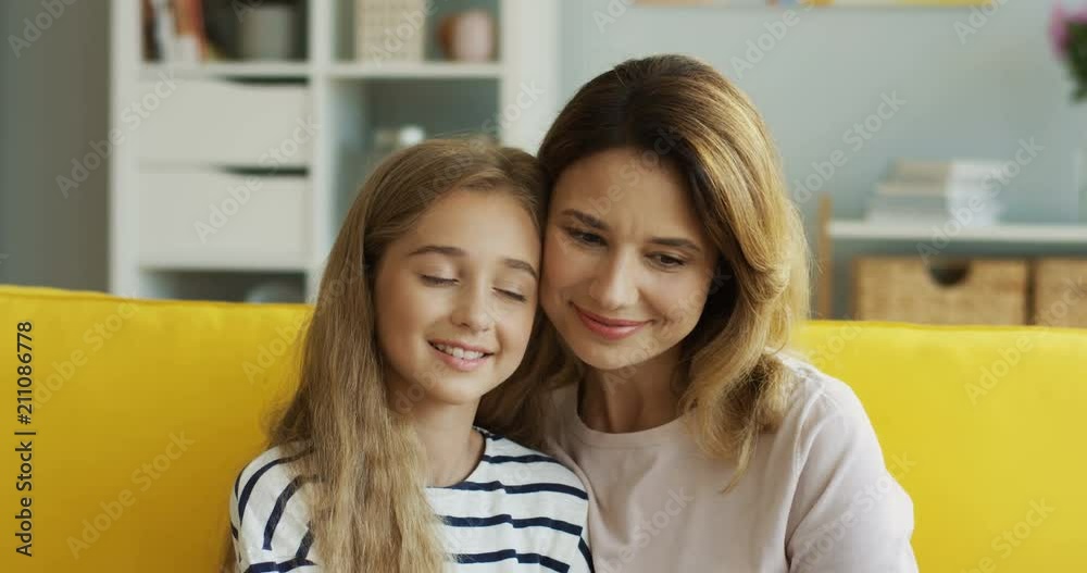 Close up of the beautiful Caucasian woman hugging her pretty teen daughter while they sitting on the couch and watching something. At home. Indoor.
