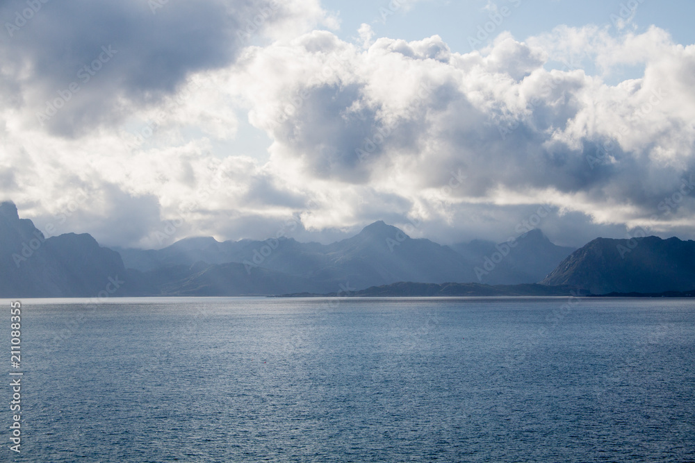 Rocky island, shoot from sea side. Mountains, deep blue sea, sun rays between heavy clouds, Norway