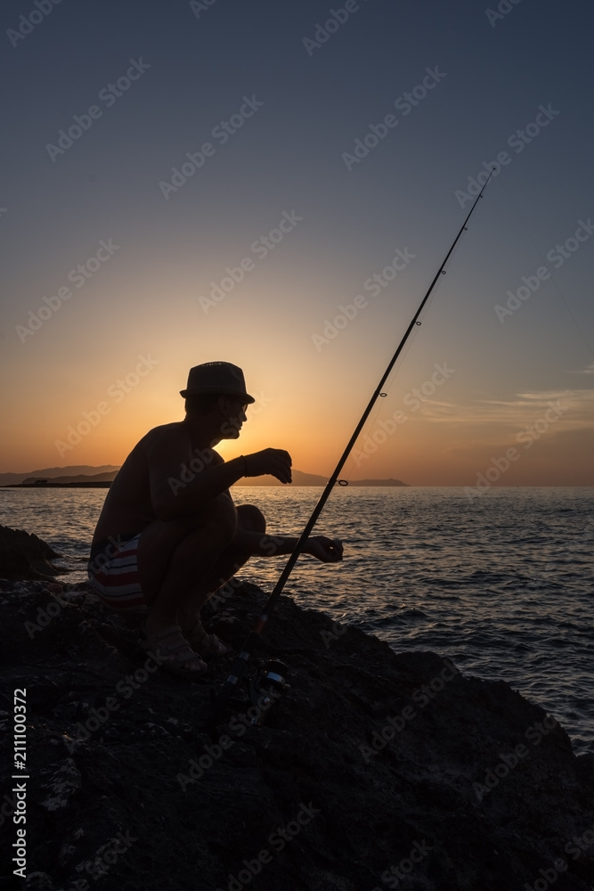 An adult man fishing on the rocks at sunset, on the seashore in Greece