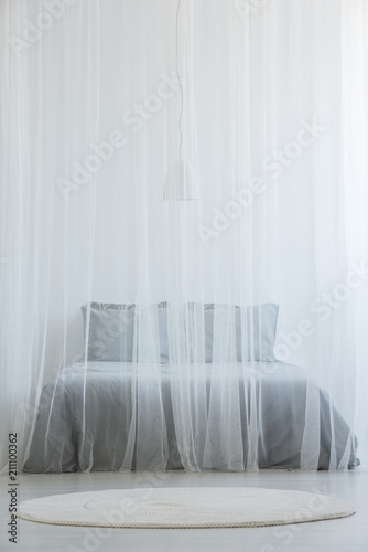 Mosquito net bed with grey bedding standing in white room interior with round rug on the floor