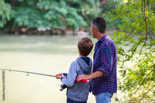 Father with son fishing at river bank, summer outdoor. Man and young boy standing at river bank with rod and fishing. Family leisure, parenting.