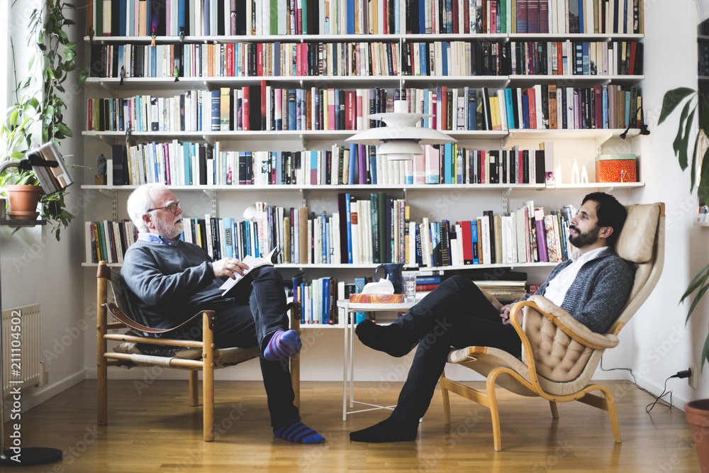 Therapist communicating with man while sitting by book shelf at home office Therapist communicating with man while sitting by book shelf at home office