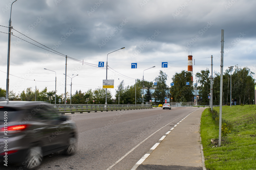 Cars drive on city road. Motion blur. Pointers and road signs on cross. Overcast. Storm rain clouds in sky.