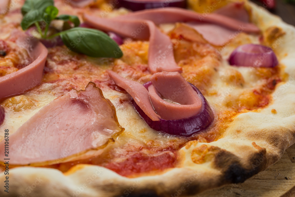 Delicious italian pizza with ham, sausages and onion decorated by basil leaves on wooden background