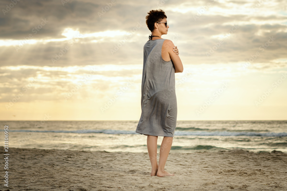 beautiful woman in a gray dress is looking at the sea on the beach at sunset beautiful woman in a gray dress is looking at the sea on the beach at sunset