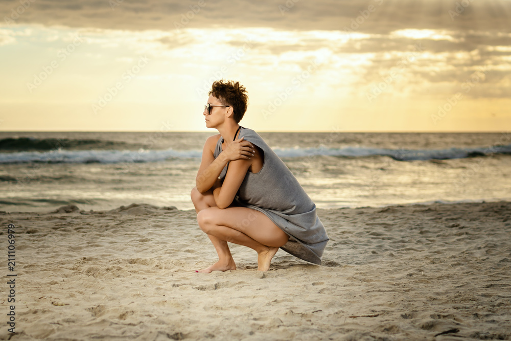 beautiful woman in a gray dress crouches on the sand at the beach at sunset beautiful woman in a gray dress crouches on the sand at the beach at sunset