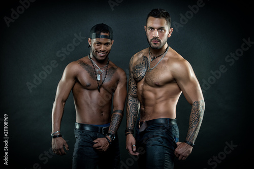 Machos with muscular tattooed torsos look attractive, dark background. Masculinity concept. Guys sportsmen with sexy muscular torsos. Athletes on confident faces with nude muscular chests