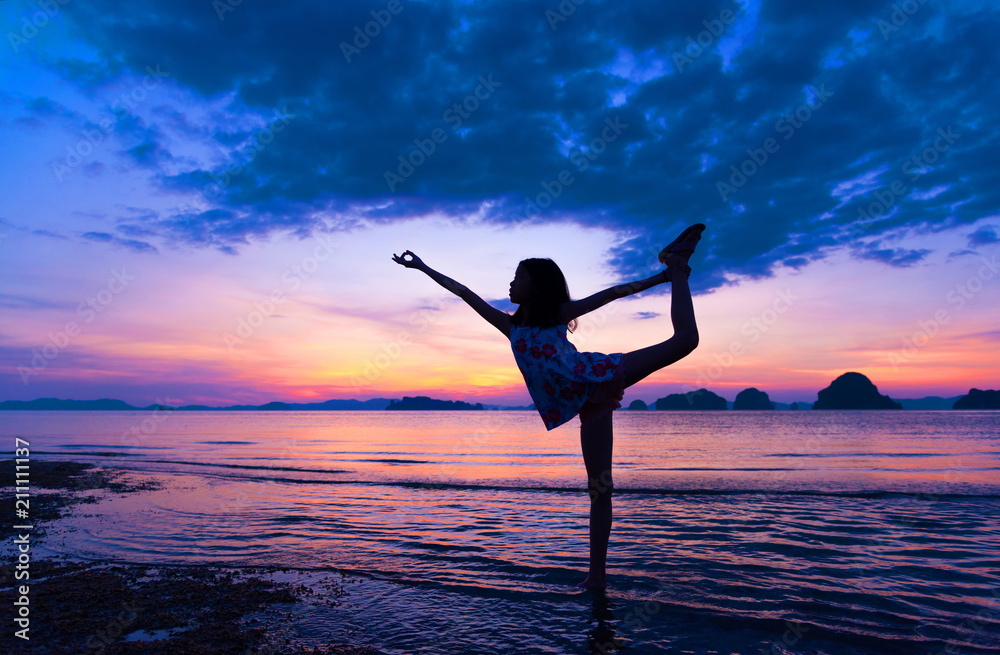 Silhouette of young girl practicing Yoga Warrior pose on tropical beach with sunset sky background. Healthy teenage woman lifestyle balanced practicing meditate and energy yoga. Healthy life Concept.