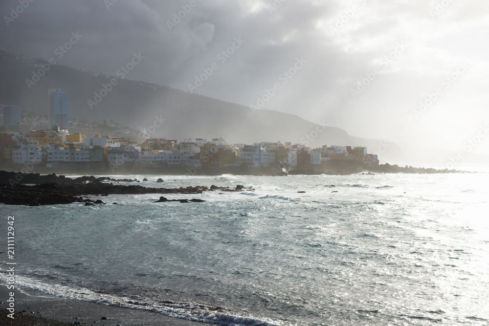 Sunrays over the coastline with houses during sunset at the black beach at Puerto de la Cruz, Tenerife, Canary Islands, Spain