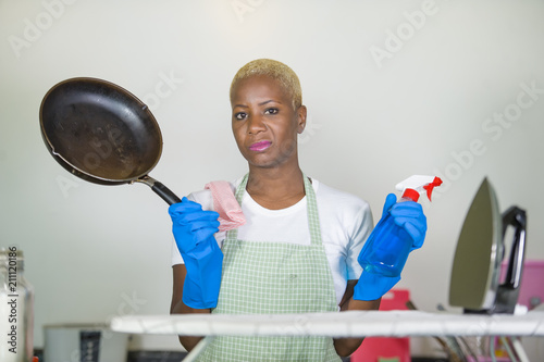 young beautiful sad and depressed afro American black woman with detergent  spry bottle and iron working upset at home kitchen overworked cleaning