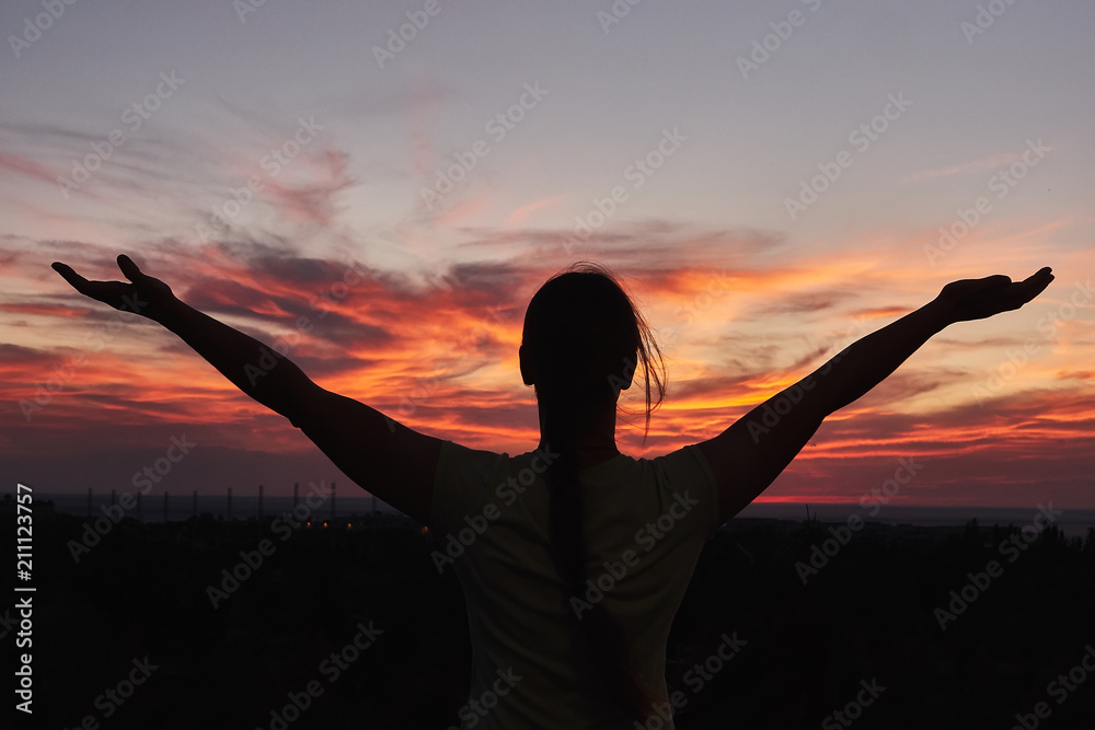 Silhouette of the woman spreading arms and enjoying breathtaking view over fields in sunset light.