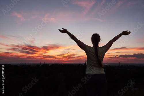 Silhouette of the woman spreading arms and enjoying breathtaking view over fields in sunset light.