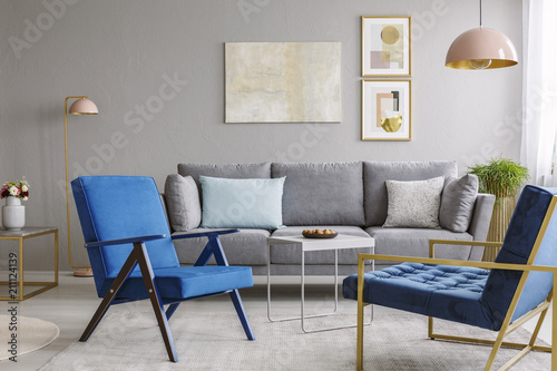 Two blue armchairs and a gray sofa standing in a living room interior with golden decorations. Real photo