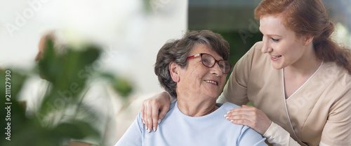 Close-up of a tender caregiver with her hands on the shoulders of a senior woman inside her home. Blurred surrounding. Panorama.