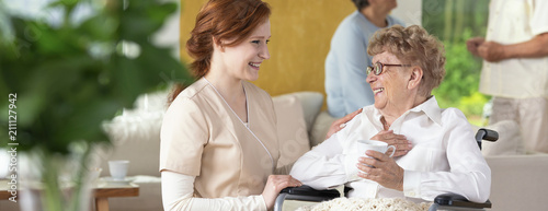 Young caretaker making a geriatric pensioner in a wheelchair laugh during leisure time in a common room of a luxury rehabilitation center. Blurred surrounding. Panorama.